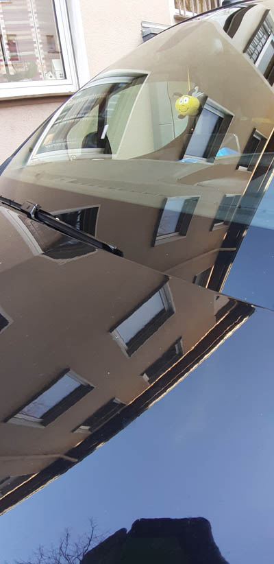 Foto-Beispiel.  In einer Autoscheibe und auf der Motorhaube  spiegelt sich der Himmel und eine Häuserzeile. Das Foto zeigt einen Ausschnitt.
Im Hintergrund ist eine Häuserreihe zu sehen.