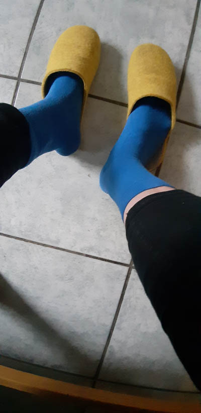 Foto-Beispiel. Zwei Füße in blauen Socken stehen auf einem hellgrauen gefliesten Küchenboden. Die Füße tragen gelbe Pantoffeln aus Filz.