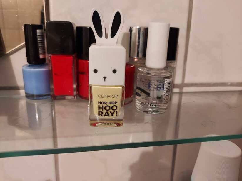 Foto-Beispiel zum Thema Osterhasen-Go! Ein hellgelber Nagellack steht auf einem Plexiglas-Stück. Das Gefäß hat eine weiße Kappe mit Hasenohren und aufgezeichnetem Gesicht. In der Nähe steht Nagellack in Rottönen und Klarlack.