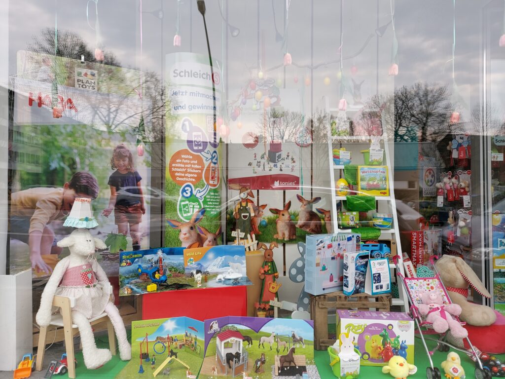 Beispiel-Foto zum Thema Osterhasen-GO im Stadtteil.Ein Schaufenster eines Spielzeugladens zur Frühlingssaison dekoriert mit vielen Figuren , einem Kiderbuggy, Küken, einer Schaaflampe und Hasen. Es sieht aus wie ein Wimmelbild.