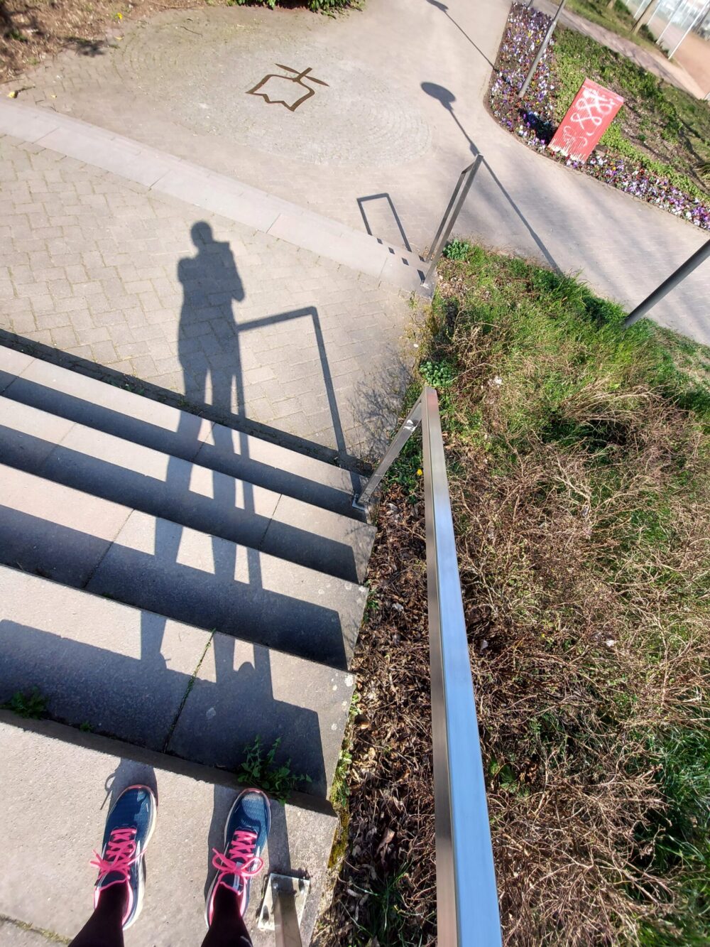 Beispiel-Foto zum Thema #Rahmen-Content im Stadtraum. Ein Geländer an einem Treppenaufgang im Außenraum wirf einen Schatten in die Sonne. Die fotografierende Person ist als Schatten zu sehen und ihre Laufschuhe mit pinkfarbenen Schnürsenkeln.