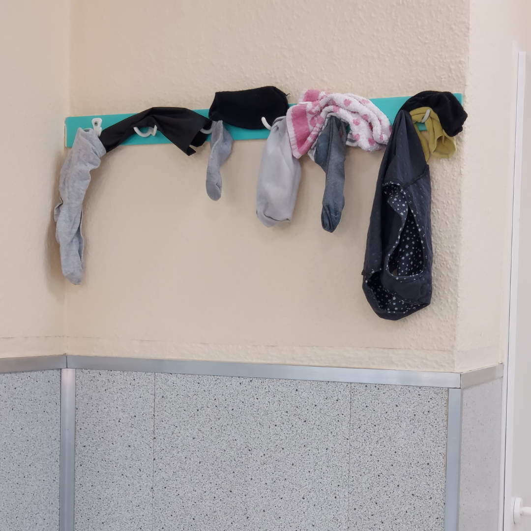 Beispiel-Foto, das Prinzip "And-on(e) im Stadtraum in einem Waschsalon, drei hellgraue und zwei schwarze Socken, ein dunkelgrauer Socken, ein weißes Handtuch mit rotem Muster und ein dunkelblaues Unterhemd mit weißen Punkten hängen an einer Hakenleiste vor einer ockergelben Wand