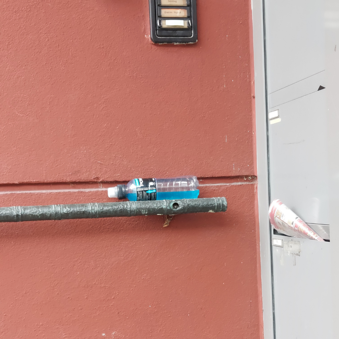 Beispiel-Foto zum Thema Add-one und Add-on. Eine Plastikflasche mit einer blauen Flüssigkeit ist waagerecht auf einem Geländer abgelegt. Das Geländer ist an einer rostroten Hauswand montiert. Aus einem Briefkastenfach an der Haustür lugt eine Zeitung heraus.