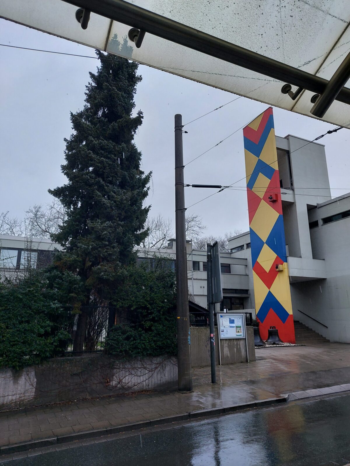 Beispiel-Foto zum Thema #Handy-Malerei. Reihe Primärfarben. Das Foto zeigt den Blick unter dem Dache einer Bahnhaltestelle an einem Regentag. Die Wand eines Gebäudes ist mit einem Rautenmuster in gelb-rot-blau gestrichen.