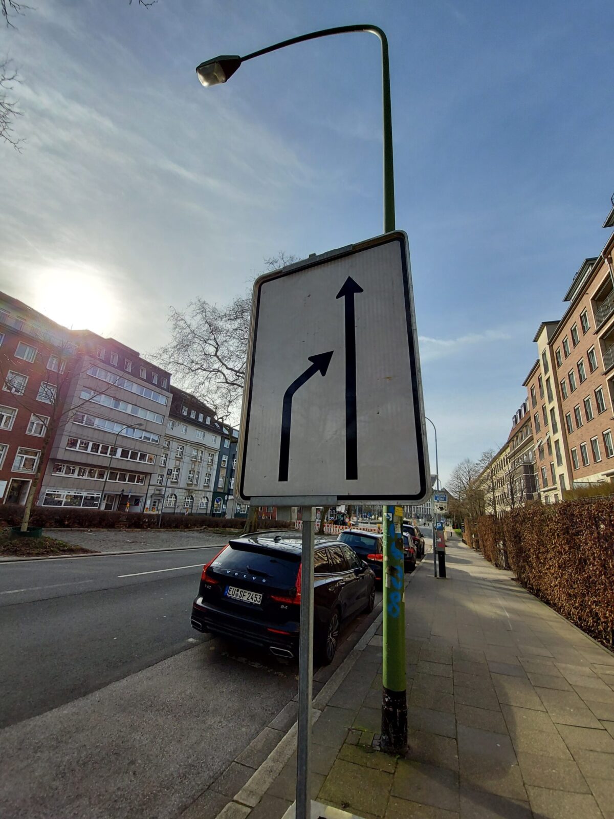 Beispiel-Foto zum Thema "Take 2" Ein Straßenschild mit zwei Pfeilen im Stadtraum im Abendlicht. Die Straßenlampe ist ausgeschaltet. Zu sehen ist ein Straßenzug und an der rechten Seite parkende Autos neben dem Gehsteig.