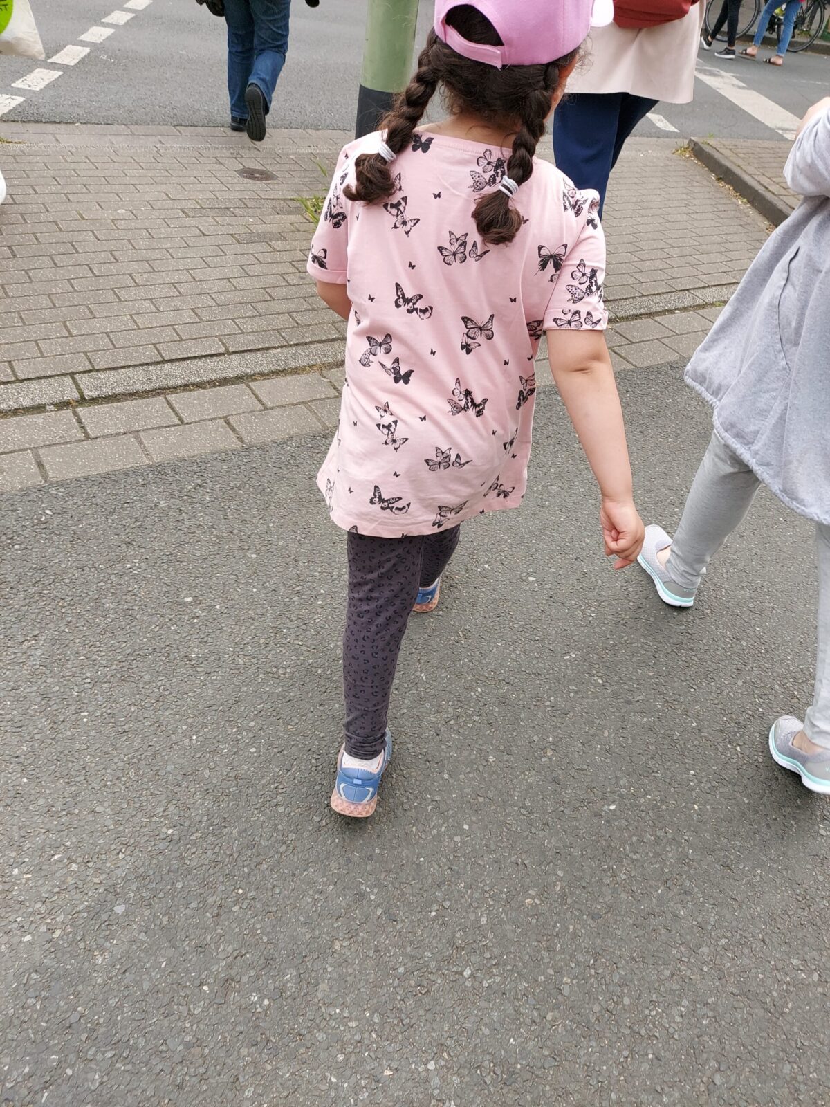 Beispiel-Foto zum Thema #cityzoo.  Ein Kind in Rückenansicht an einer Straßenkreuzung. Das Kind trägt ein rosafarbenes T-Shirt mit Schmetterlingen., blaue Schuhe, eine rosafarbenen Basecap. Die dunklen Haare sind zu zwei Zöpfen geflochten. 