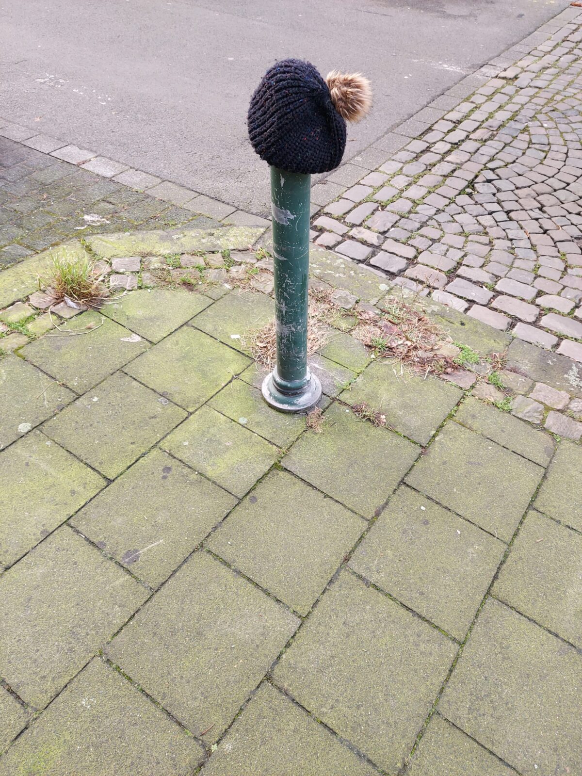 Beispiel-Foto zum Starterkit Sockel(it)up! im Stadtraum, eine Säule mit blauer Wollmütze mit Bommel an der Straßenecke, der Boden teilt sich in Asphalt, Pflastersteine und Platten