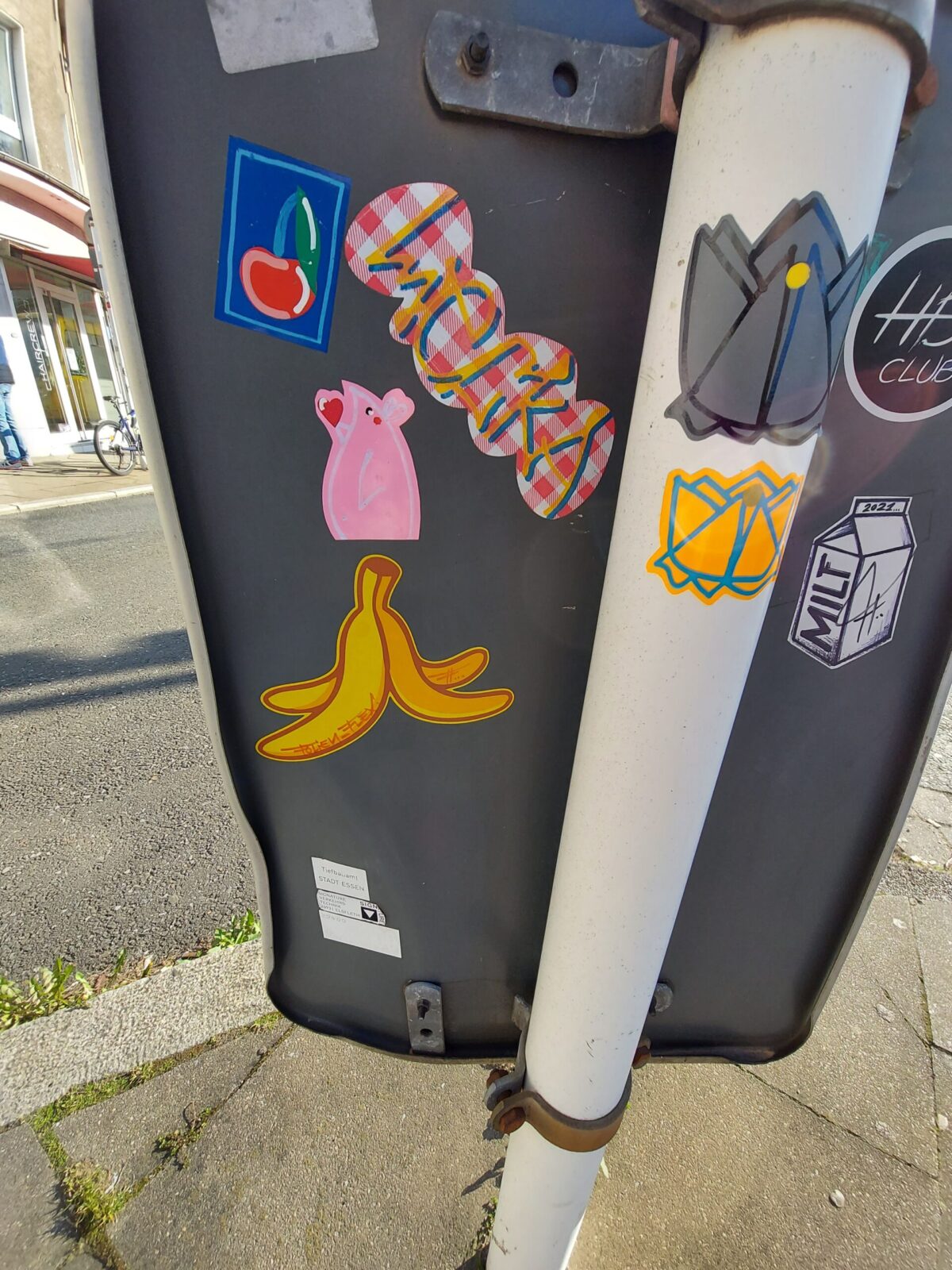 Beispiel-Foto zum Thema #cityzoo. Zu sehen ist die Rückseite eines Straßenschildes mit verschiedenen Stickern. Eine Kirsche vor blauem Hintergrund, eine Banananeschale, 2 Fledermausfiguren, ein Schriftzug von kariertem Hintergrund, ein runder Sticker eines Clubs, eine Milchtüte mit dem Schriftzug MILF
