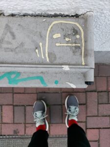 Beispiel-Foto zum Starterkit  #cityzoo. Eine gelbe Linienzeichnung auf einem grauen Stromkasten. Das Foto ist aus der Vogelperspektive aufgenommen und zeigt die Beine und roten Socken und grauen Turnschuhe mit hellblauen Schnürsenkeln der fotografierenden Person.