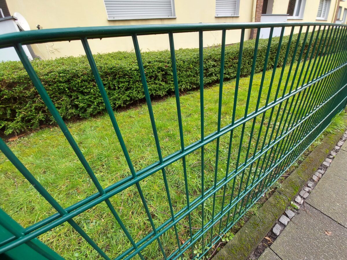 Beispiel-Foto zum Thema "Little Stages" im Stadtraum, ein grüner Gitterzaun vor einer Wiese, im HInergund eine hellgelbe Hauswand, die Fensterläden des Hauses sind geschlossen, die Hecke davor ist