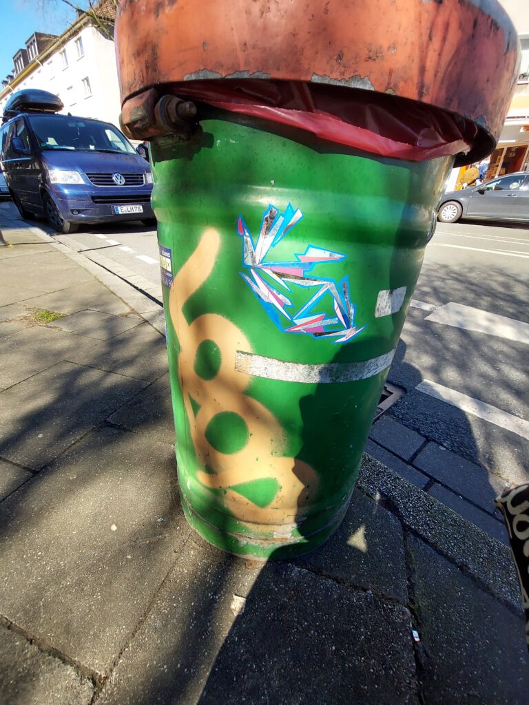 Beispiel-Foto zum Starterkit  #cityzoo. Ein glitzernder Fisch in Türkis- und Pinktönen  klebt auf einer Mülltonne. Die Mülltonne hat einen grünen Korpus und einen roten Rand. Im Hintergrund ein Straßenzug und ein blaues Auto