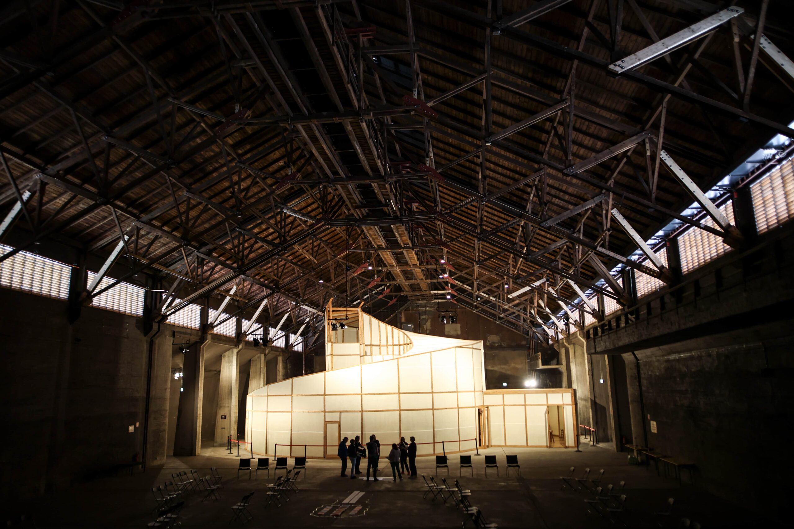 Situationen in denen Menschen ihre Ideen und Perspektiven finden. Workshop "Du und D-eine Gegenwart". Maßkonzetion für die Stiftung Zollverein durch Studio Wundermaterial. Blick in die Halle des Salzlagers auf Zollverein in Essen. Die Workshop-Teilnehmer:innen stehen mittig vor dem erleuchteten Kunstwerk "Palace of Projects". Es handelt sich um eine spiralförmige Konstruktion aus Holz, das mit einem Gewebe bespannt wurde. Durch das Gegenlicht sind die Personen als Schattenrisse zu sehen. Links und rechts der Gruppe stehen jeweils vier Stühle.