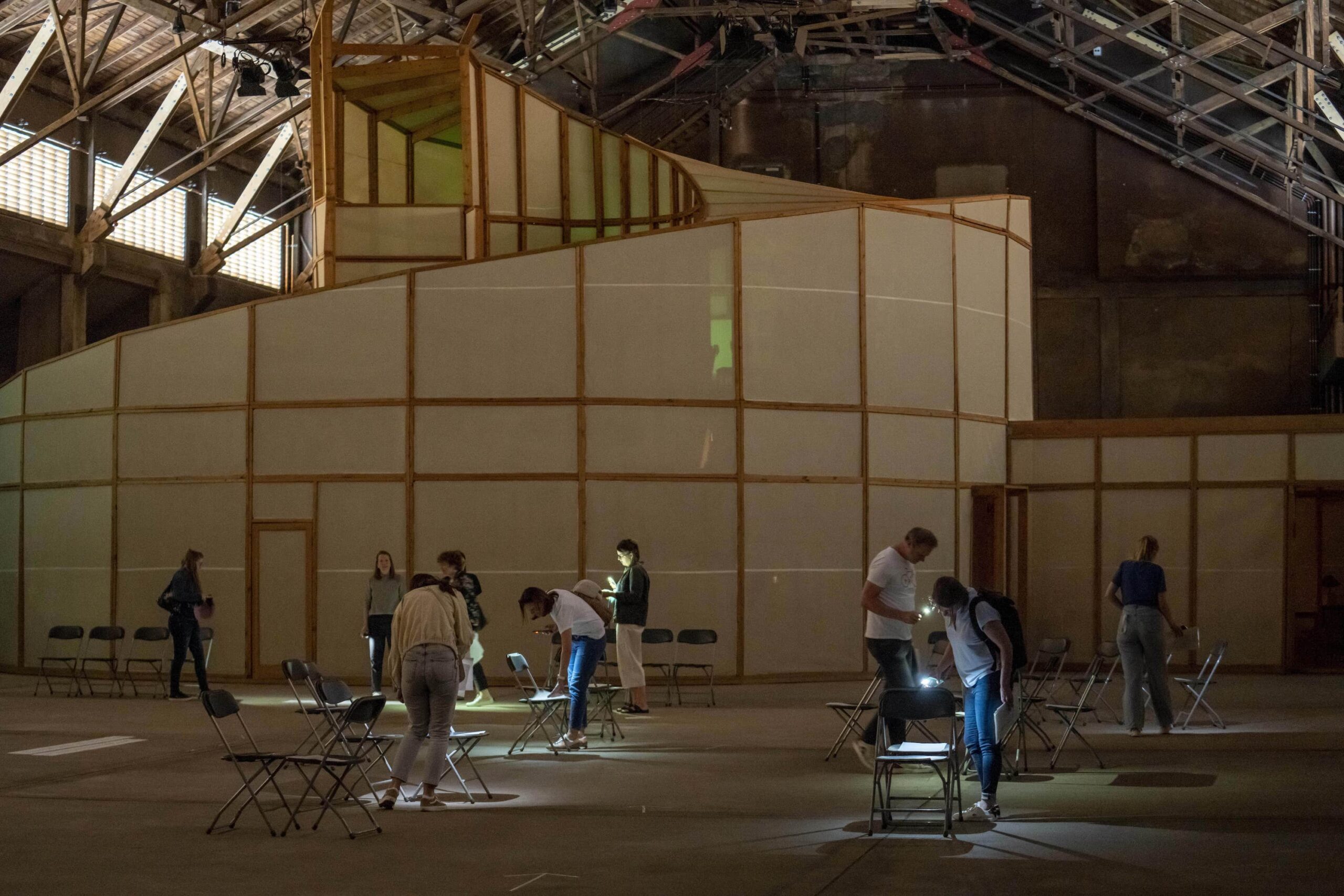 Fragen und Ideen finden und zusammen ins denken kommen durch Kunst. Maßkonzeption für die Stiftung Zollverein. "Suchende Lichter" im Workshop. Insgesamt sechs Personen bewegen sich vor dem Kunstwerk "Palace of Projects" von Ilya und Emilia Kabakov durch den Raum. Sie leuchten mit ihren Handytaschenlampen und erhellen damit Zettel, die auf Stühlen platziert sind. Auf dem Boden und am Kunstwerk selbst sind mehrere Lichtreflexe zu sehen. Die Personen tragen Kleidung mit kurzen Armen und lange Hosen.